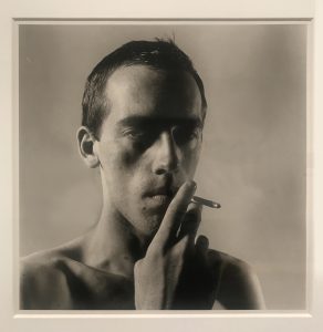 A black and white portrait photograph of David Wojnarowicz drawing on a cigarette looking at the camera