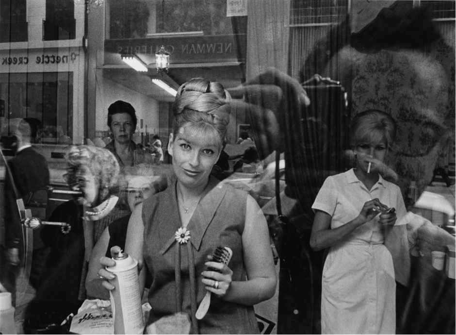 A black and white photograph of a woman looking out at the camera through the front window of a hair salon