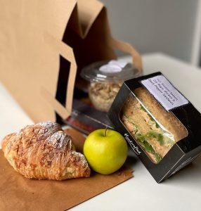 Picture of a paper bag with a croissant, apple, sandwich, and a breakfast pot.