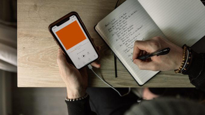 One hand holds a phone playing a recording through headphones. The other hand writes in a notebook