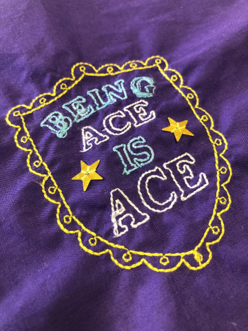 A piece of purple fabric, covered in embroidery. There is a yellow shield, with 'being ACE is ACE' stitched into the middle, with a yellow golden star either side.