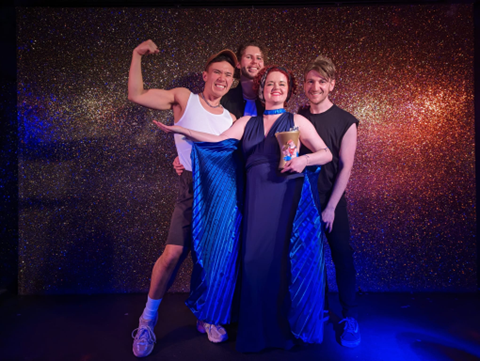 A group of 4 people stood in a group against a sparkling wall under dim lighting. Rosanna is stood at the front of the group wearing a long blue floor length gown. 