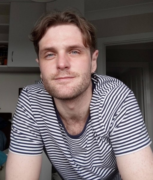 A portrait of Andrew Hall. He is wearing a tshirt with black and white stripes and has brown hair. 