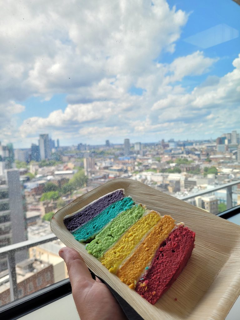 A large wedge of pride themes layer cake held up against a window with views of South London in the background.