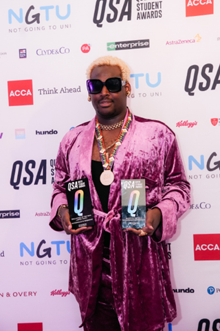 Taj at the Queer Student Awards holding two trophies featuring a big Q on each. Taj is wearing a plum velvet 2 piece outfit, a medal around his neck and stylish sunglasses. 