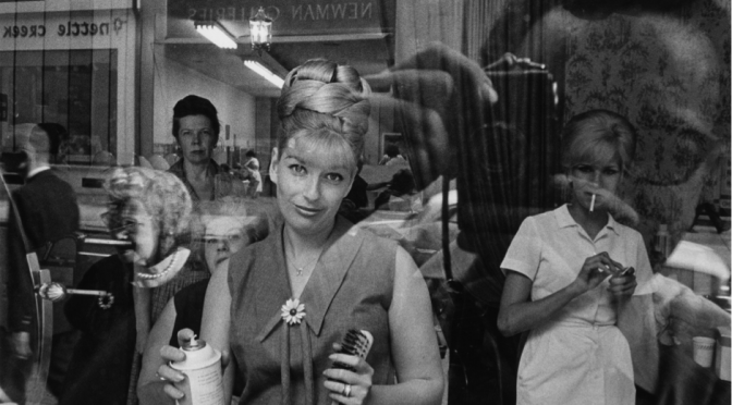 A black and white photograph of a woman looking out at the camera through the front window of a hair salon