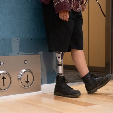 Lower body crop of an Indigenous person with a prothestic leg standing next to an open elevator and its floor-located call buttons.