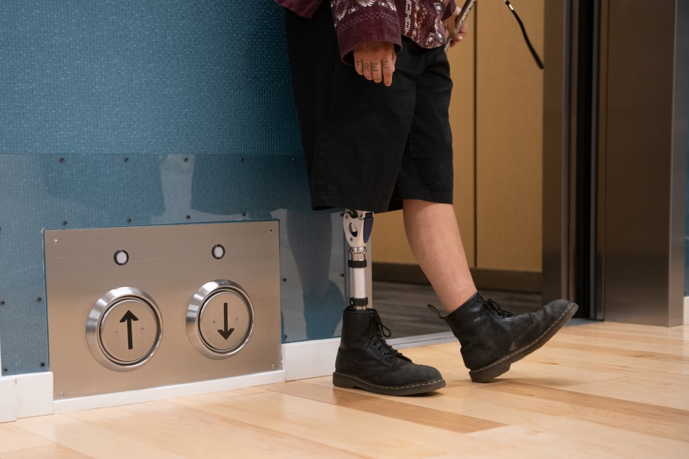 Lower body crop of an Indigenous person with a prothestic leg standing next to an open elevator and its floor-located call buttons. 