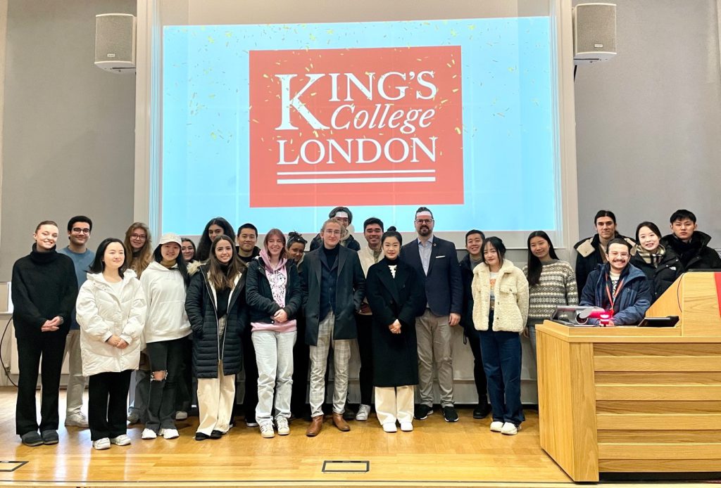 Around 25 International Marketing students and staff pose for a photo on a stage under the King's College London logo